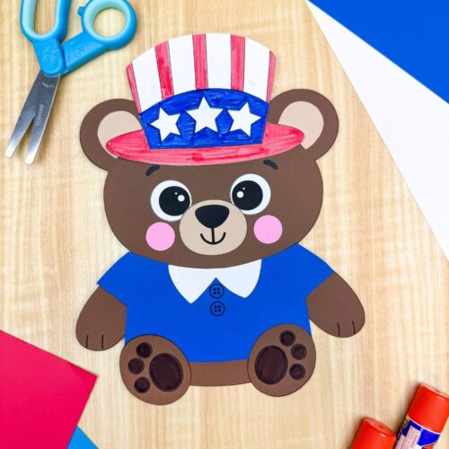 A completed patriotic teddy bear paper craft with a blue shirt and Uncle Sam hat surrounded by scissors, glue sticks, and colorful craft paper