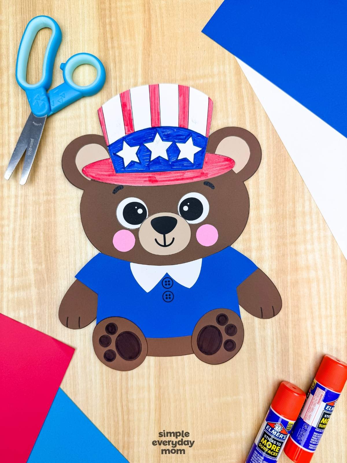A completed patriotic teddy bear paper craft with a blue shirt and Uncle Sam hat surrounded by scissors, glue sticks, and colorful craft paper
