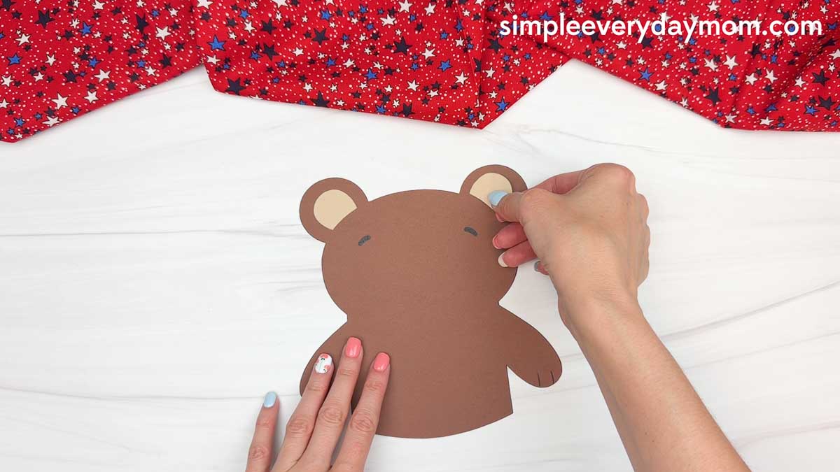 Hands attaching the teddy bear’s ears to a brown paper bear craft for a patriotic teddy bear activity
