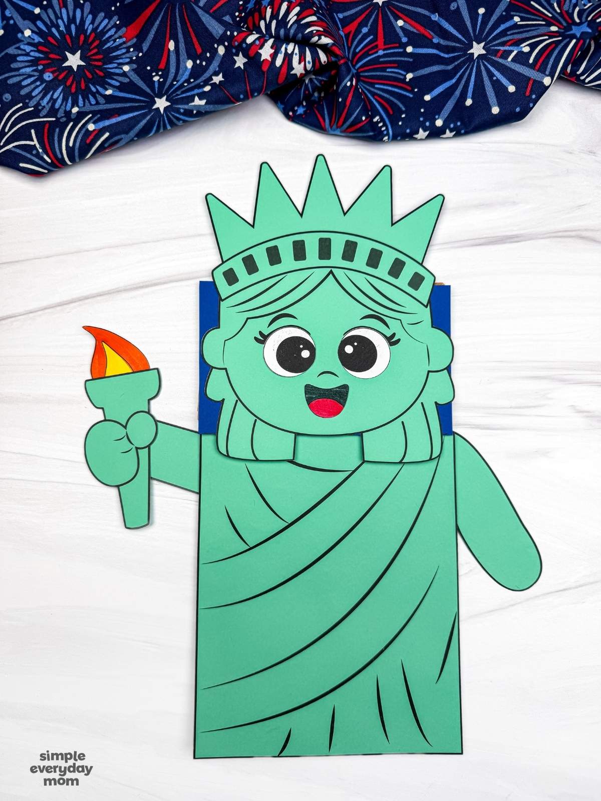 A completed craft showing a Statue of Liberty paper bag puppet with a torch and smiling face on a white tabletop.