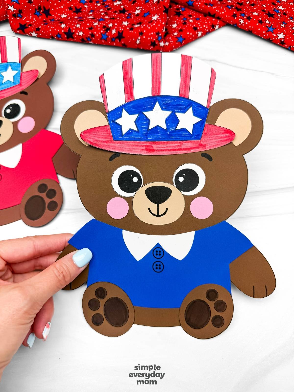 Hands holding a completed patriotic teddy bear paper craft with a blue shirt and Uncle Sam hat for a Fourth of July kids activity