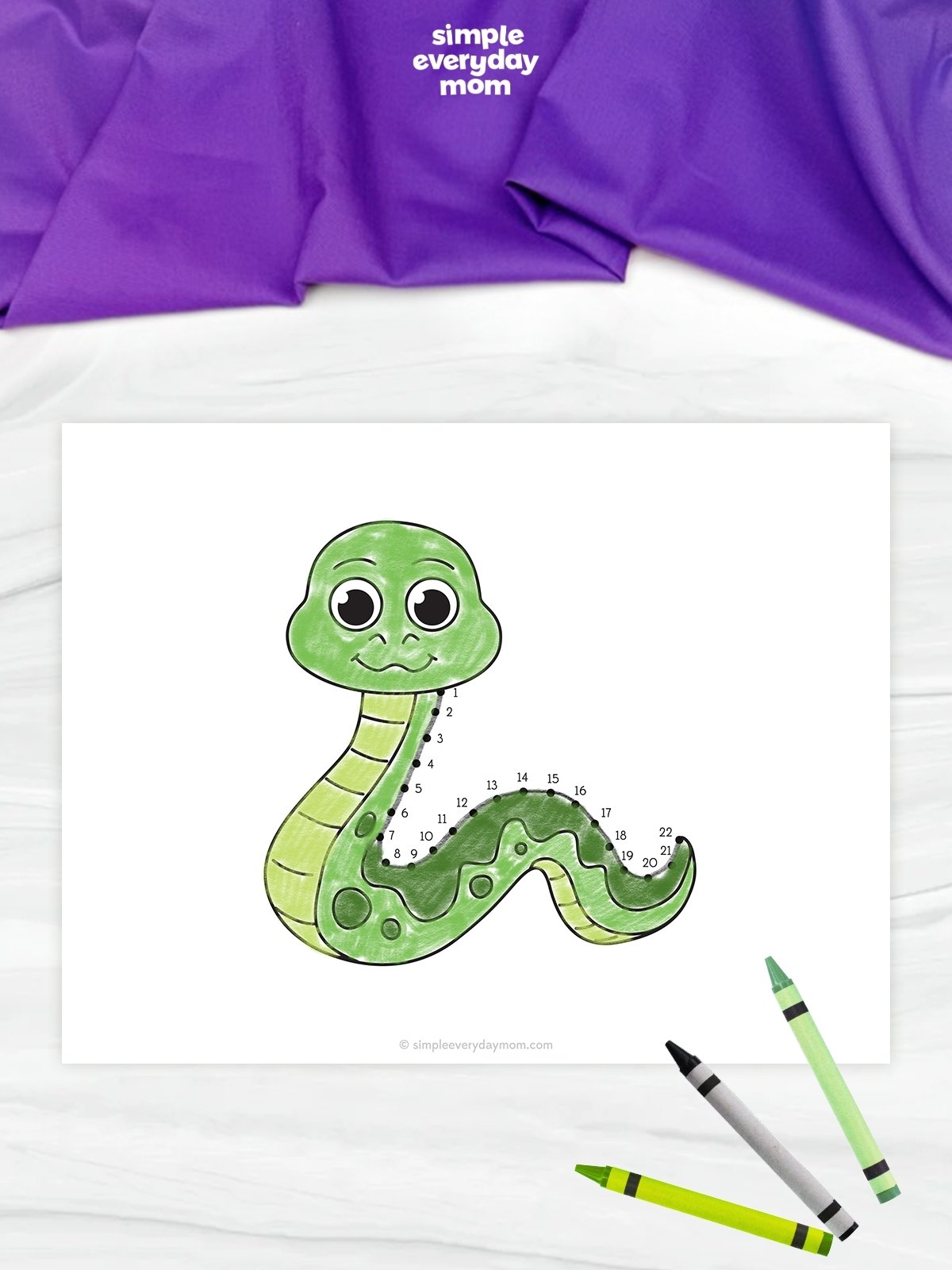 This image features a completed snake connect-the-dots worksheet placed on a light marble-like surface. The smiling green snake is colored with different shades of green, and numbered dots trace part of its body from head to tail. Three crayons are positioned in the lower-right corner, while a purple fabric and the Simple Everyday Mom logo appear at the top of the image.