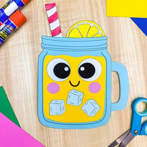 A completed lemonade paper craft with a smiling mason jar, striped straw, lemon slice, and craft supplies on a wooden table.
