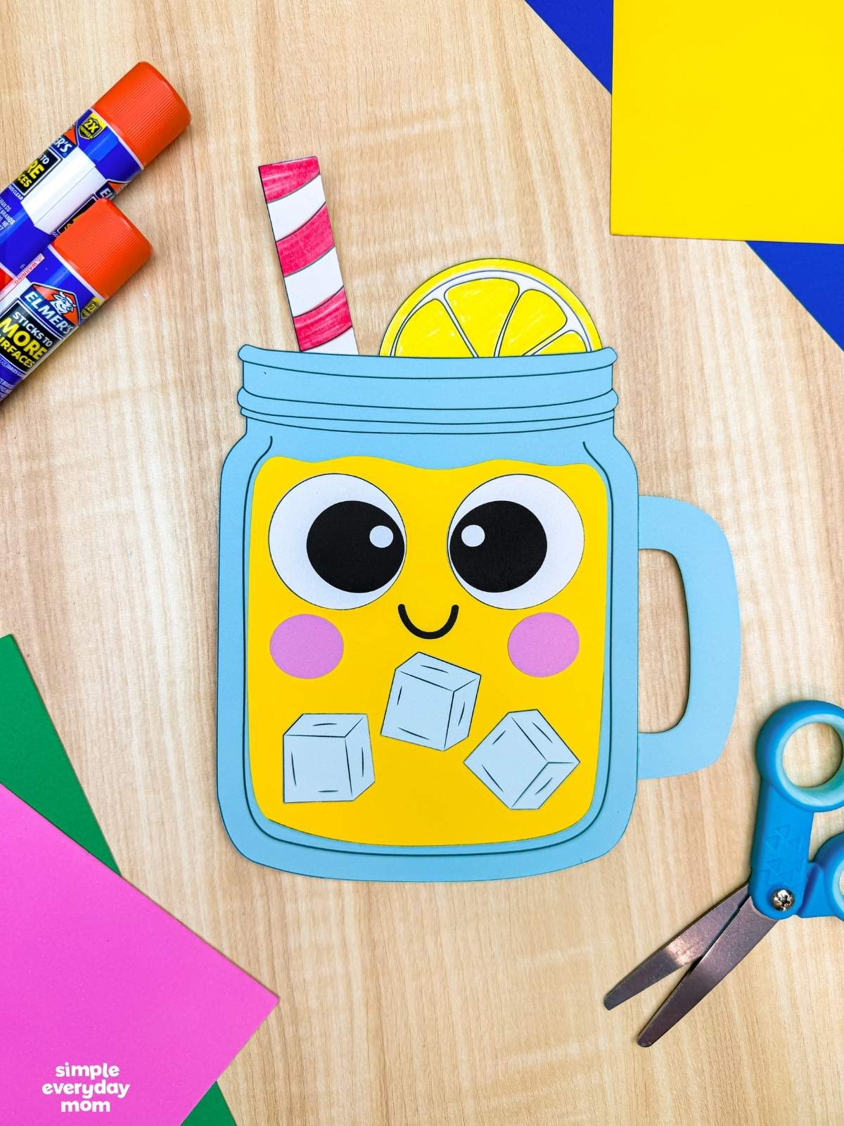 A completed lemonade paper craft with a smiling mason jar, striped straw, lemon slice, and craft supplies on a wooden table.