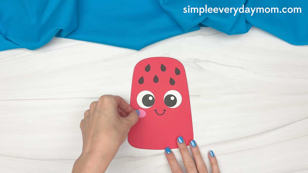 Hands placing a pink cheek piece onto a completed summer popsicle craft with a smiling face and watermelon seed details.