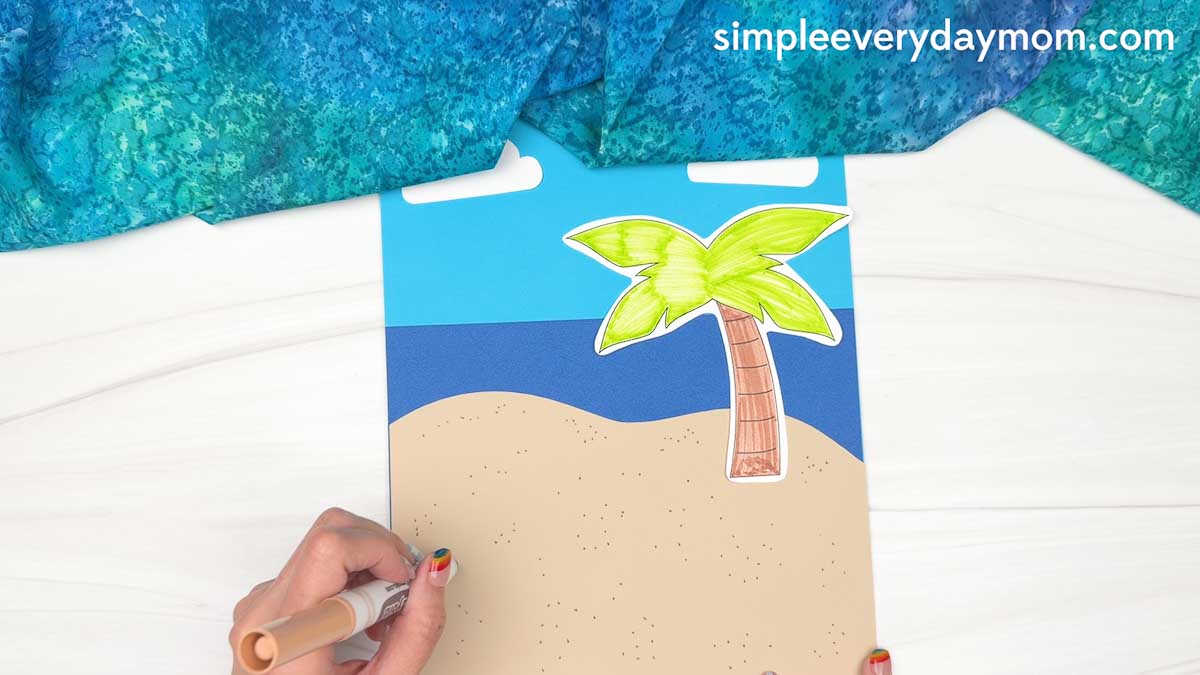 Hands adding sand texture details with a marker to a beach scene background for a reindeer summer craft.