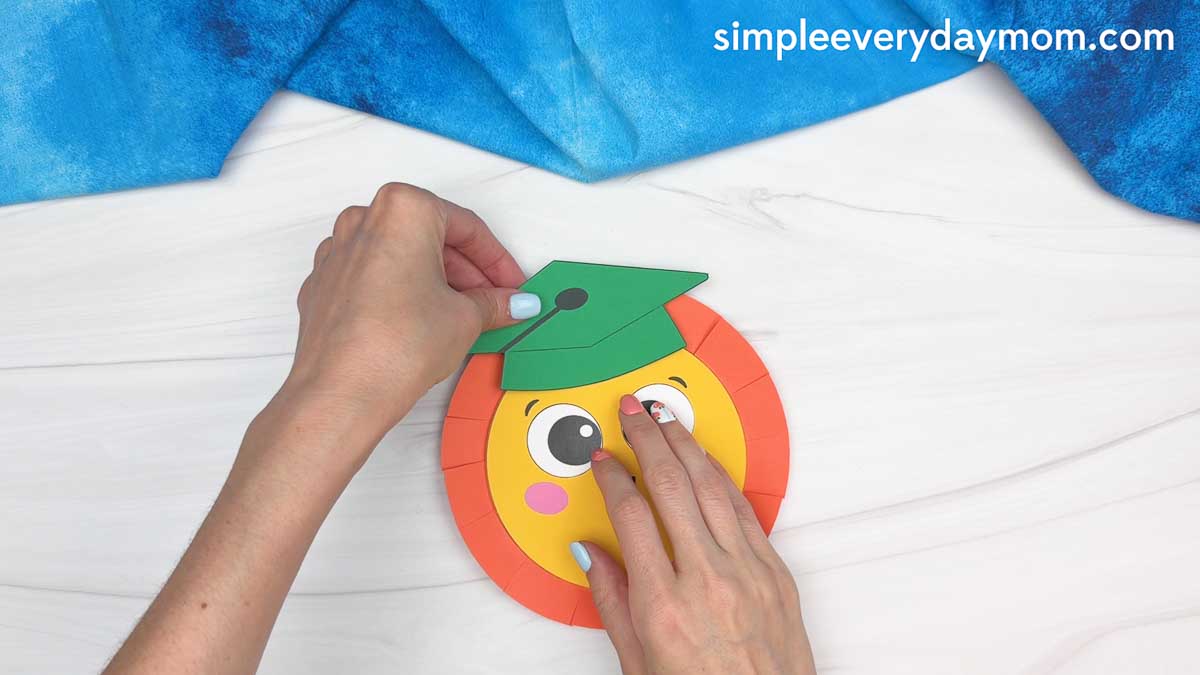 Hands attaching a green graduation cap to a smiling sun craft with folded orange paper rays