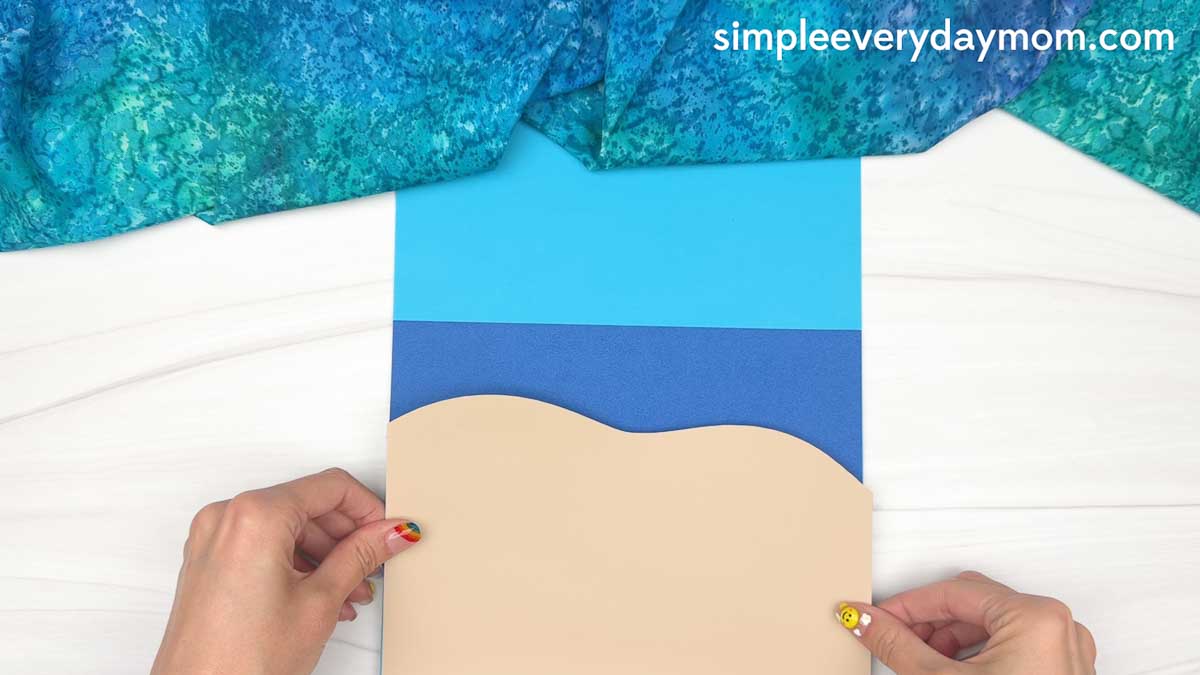 Hands positioning a tan sand-shaped paper cutout onto a blue beach background for a reindeer summer craft.