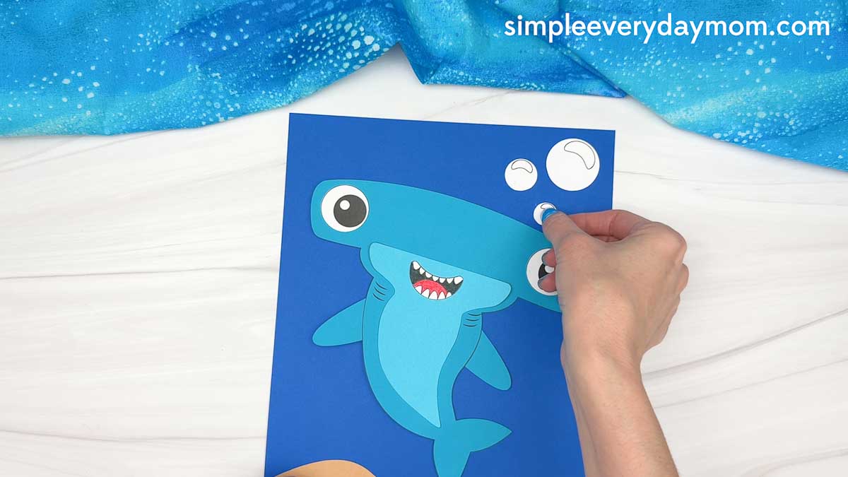 Hands attaching a paper bubble to a hammerhead shark craft on an underwater ocean background.