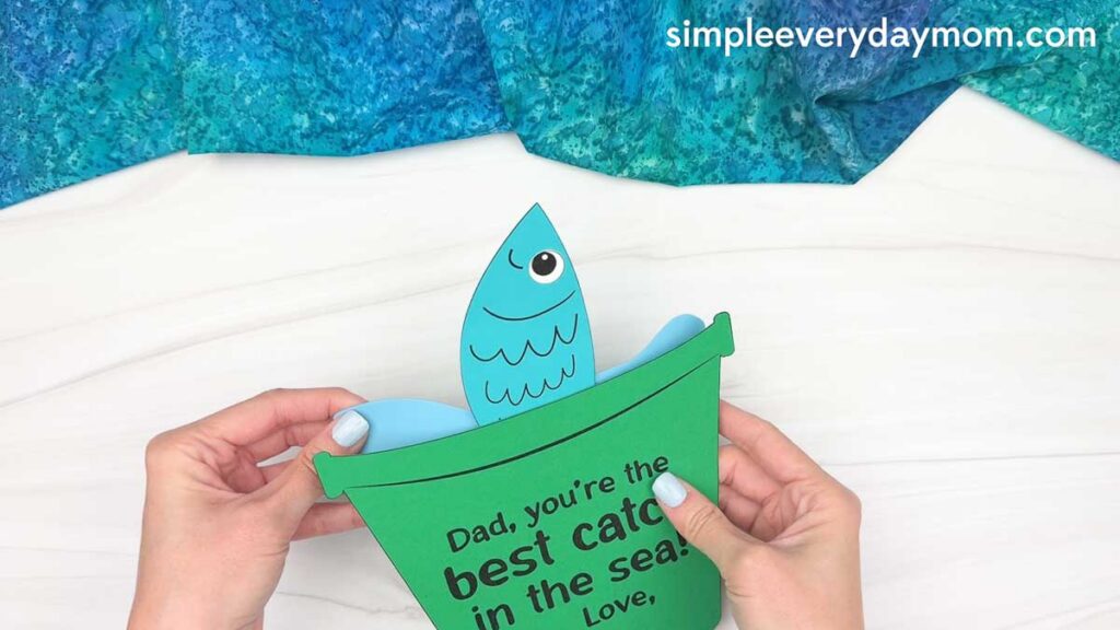 Hands holding a completed Father’s Day fishing bucket craft with paper fish and a best catch in the sea message.