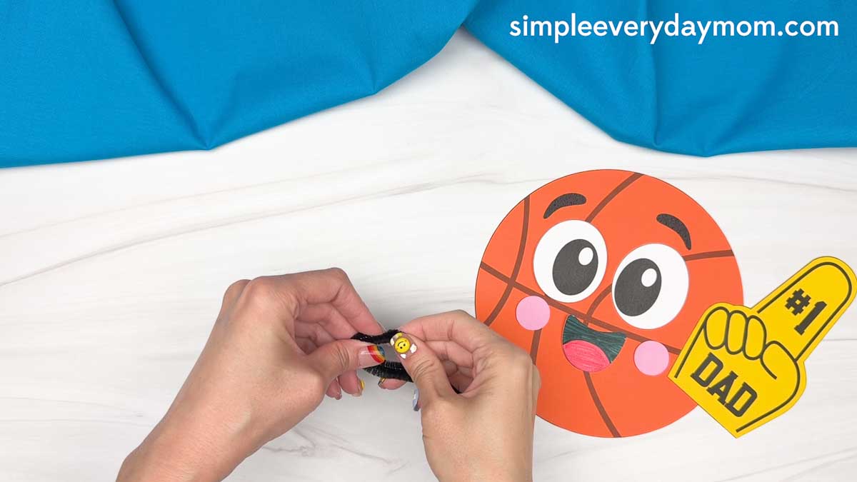 Hands attaching pipe cleaner arms to a basketball Father’s Day paper craft with a number one dad foam finger.