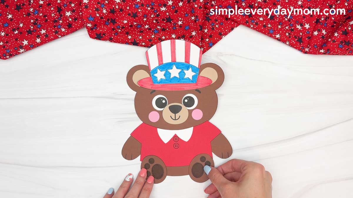 Hands attaching the teddy bear paw pieces to a completed patriotic teddy bear paper craft activity