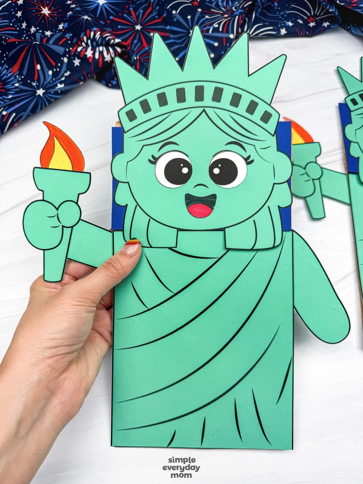 Hands holding a completed Statue of Liberty paper bag puppet craft with a torch and smiling face.