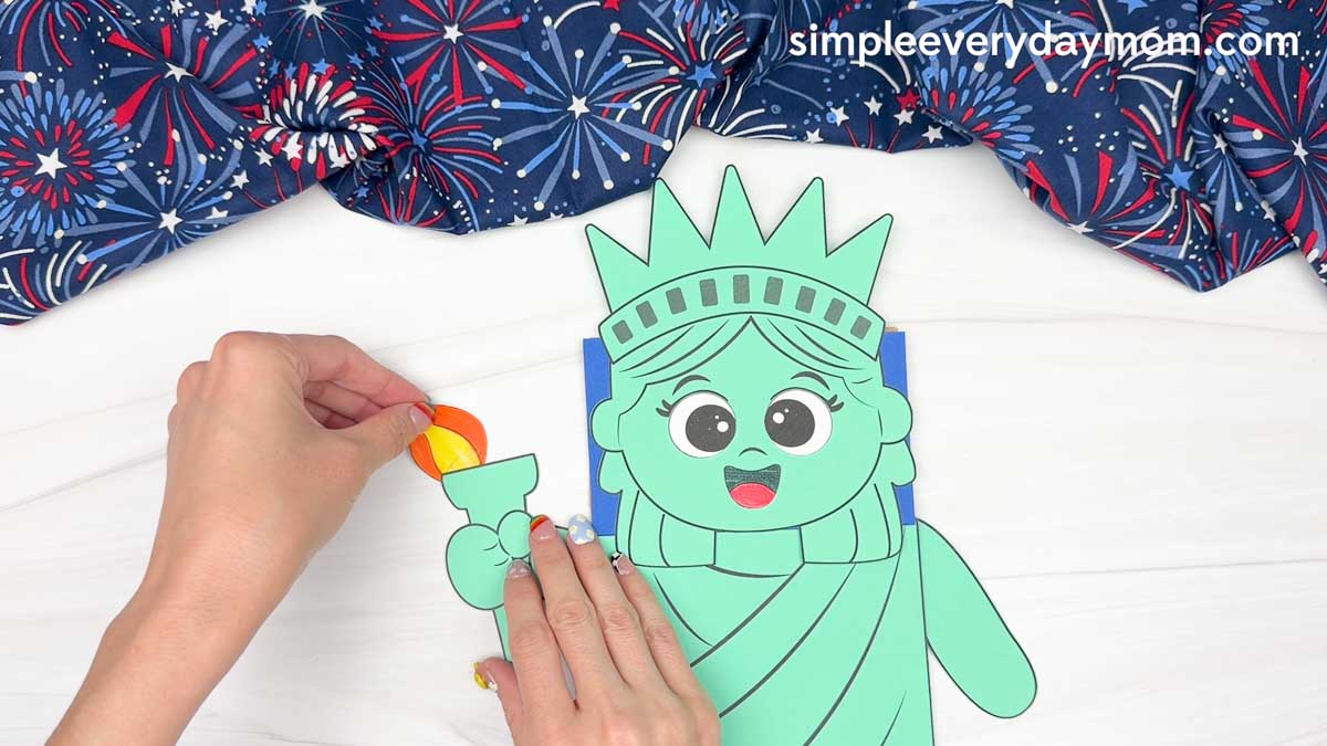 Hands attaching the flame cutout to the Statue of Liberty torch on the paper bag puppet craft.