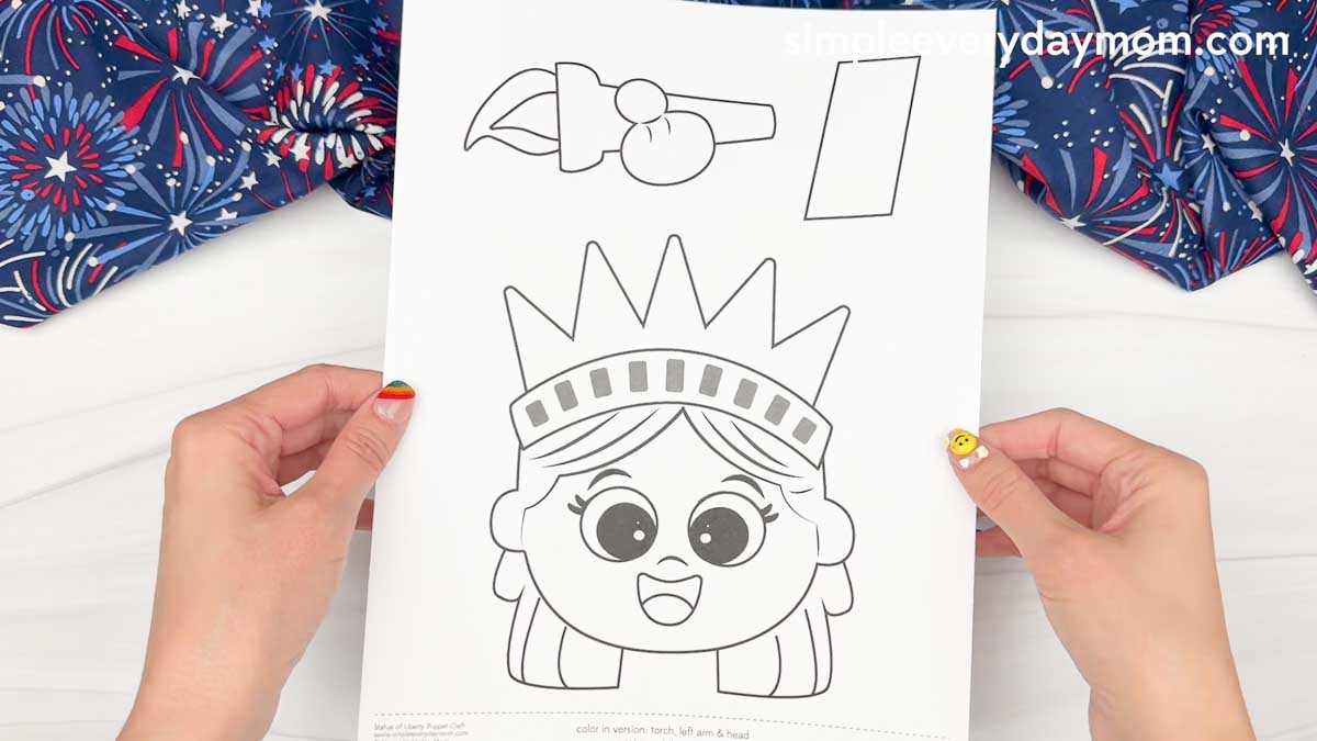 Hands holding a Statue of Liberty puppet craft template with a cartoon face and cutout pieces for the torch, arm, and crown.