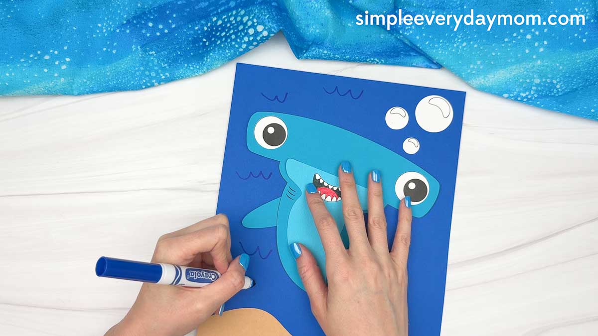 Hands drawing ocean waves around a hammerhead shark craft on a blue underwater background.