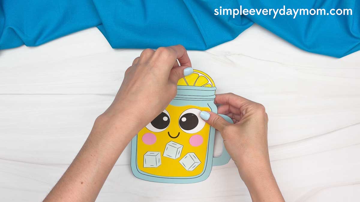 Hands placing a lemon slice on top of a completed lemonade jar craft with paper ice cubes and a smiling face.
