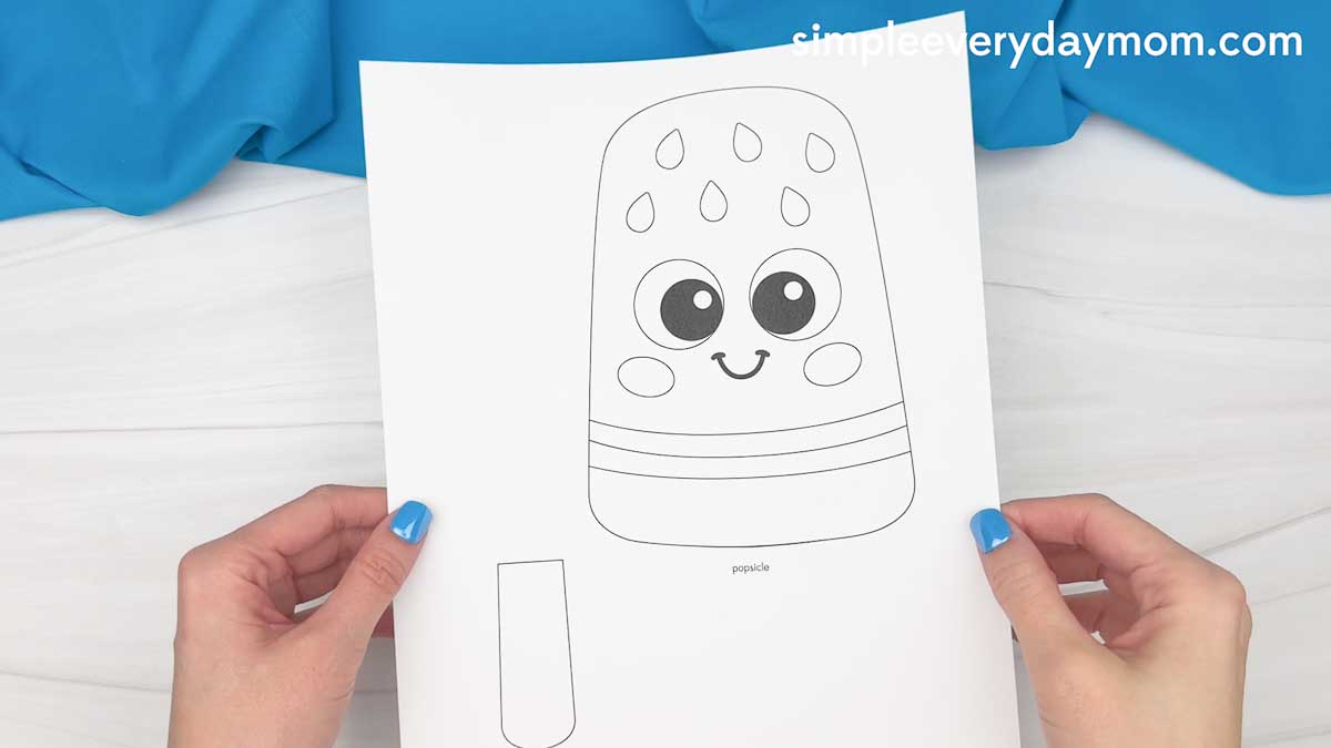 Hands holding a printable summer popsicle craft template with a smiling popsicle character and popsicle stick cutout.