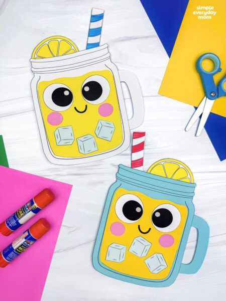 A completed lemonade paper craft with smiling mason jars, colorful paper, glue sticks, and scissors on a table.