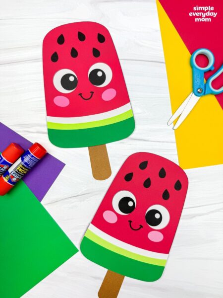 A completed craft showing two smiling watermelon popsicle crafts surrounded by colorful paper, scissors, and glue sticks.