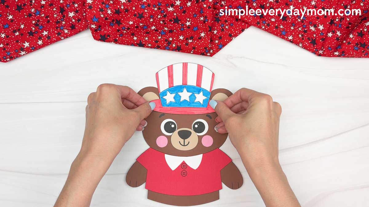 Hands placing a patriotic Uncle Sam hat onto a brown teddy bear paper craft activity