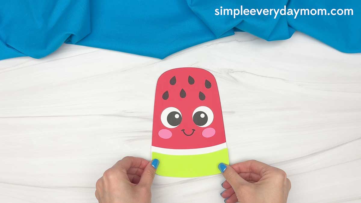 Hands holding a completed summer popsicle craft with a smiling watermelon design and bright green bottom stripe.