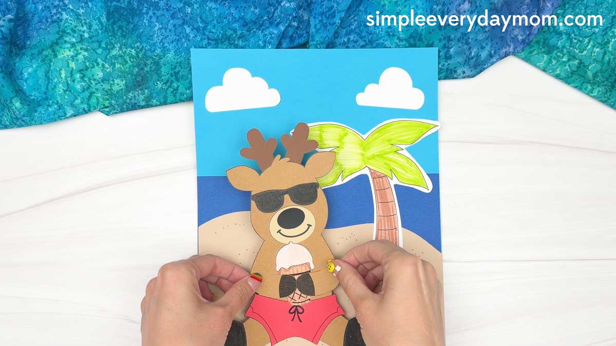 Hands positioning the completed reindeer summer craft on a beach scene background with a palm tree, clouds, and ocean.