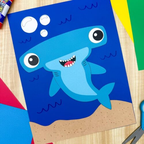 A completed hammerhead shark craft on a blue underwater background surrounded by colorful paper, scissors, and a glue stick.