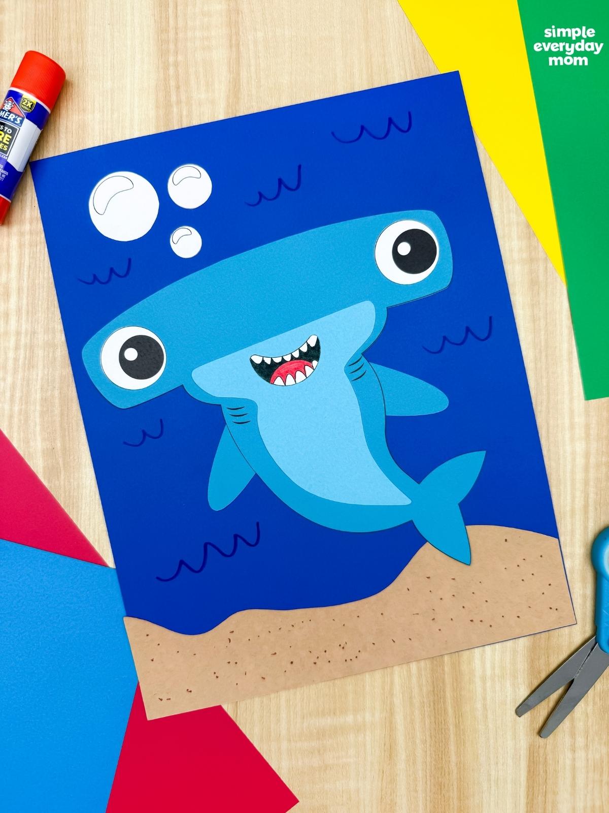 A completed hammerhead shark craft on a blue underwater background surrounded by colorful paper, scissors, and a glue stick.