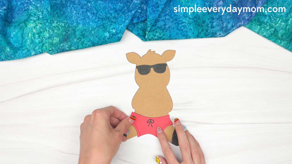 Hands attaching pink swim trunks to the reindeer body cutout for a reindeer summer craft.