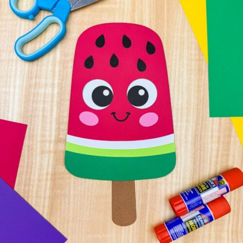 A completed craft showing a smiling watermelon popsicle craft surrounded by colorful paper, scissors, and glue sticks on a wooden surface.