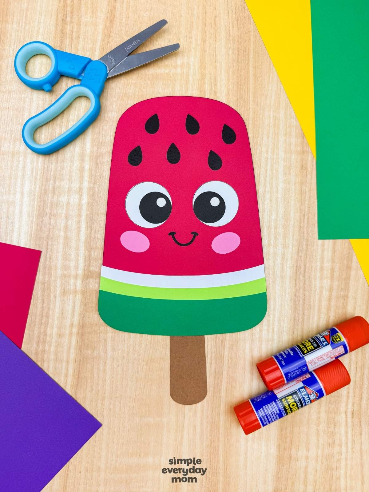A completed craft showing a smiling watermelon popsicle craft surrounded by colorful paper, scissors, and glue sticks on a wooden surface.
