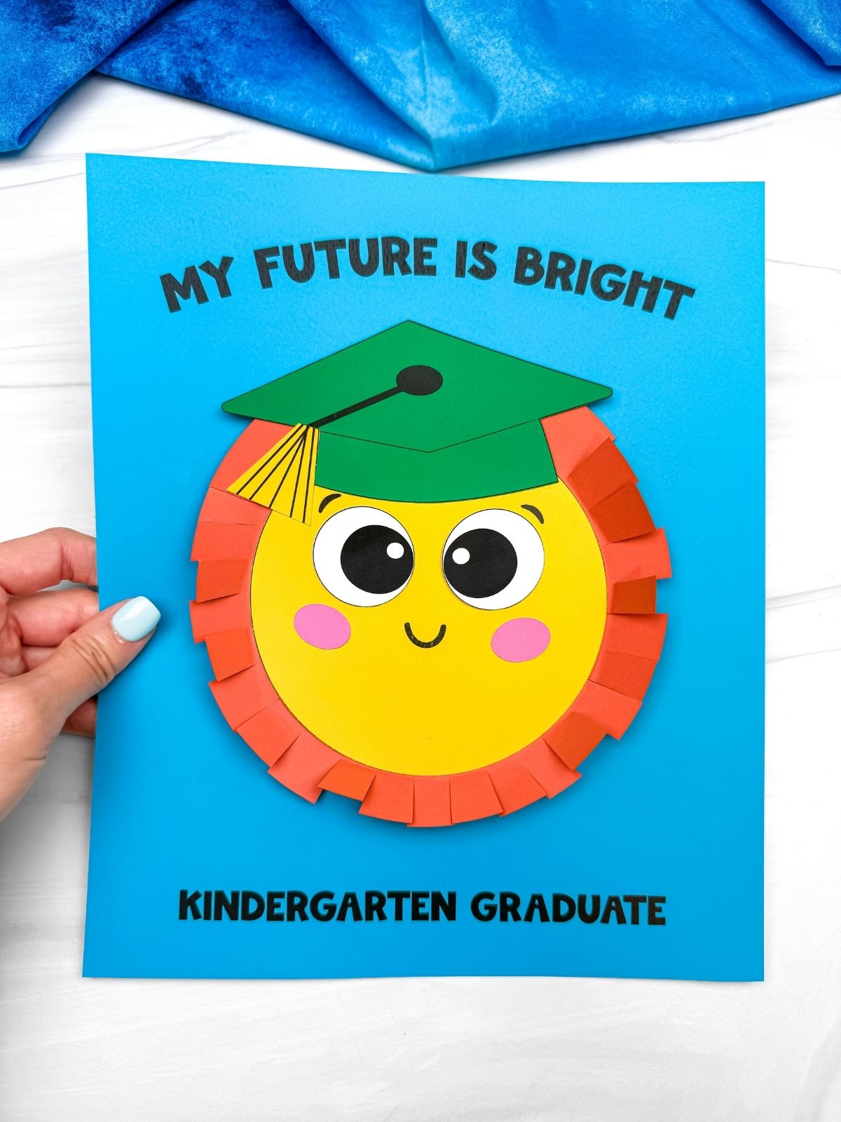 Hands holding a completed sun graduation craft card with a smiling sun wearing a green graduation cap and “My Future Is Bright” text on blue paper