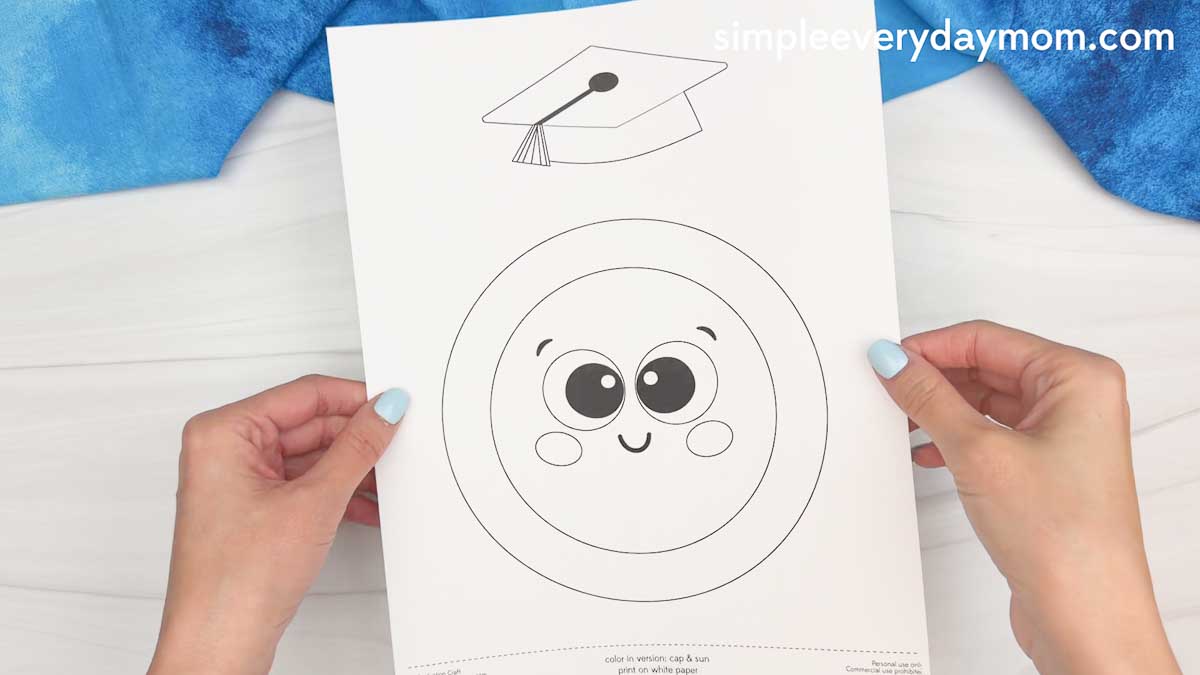 Hands holding a printable sun graduation craft template with a smiling sun face and graduation cap outline on white paper