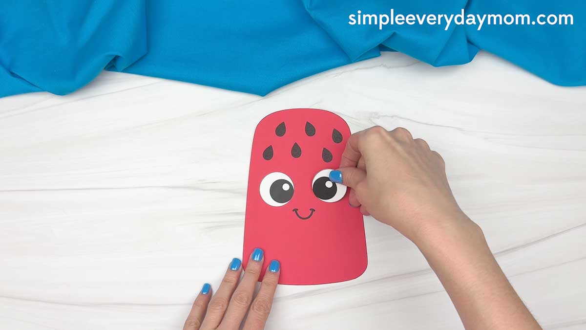 Hands attaching a large googly eye to a completed summer popsicle craft with watermelon seed details and a smiling face.