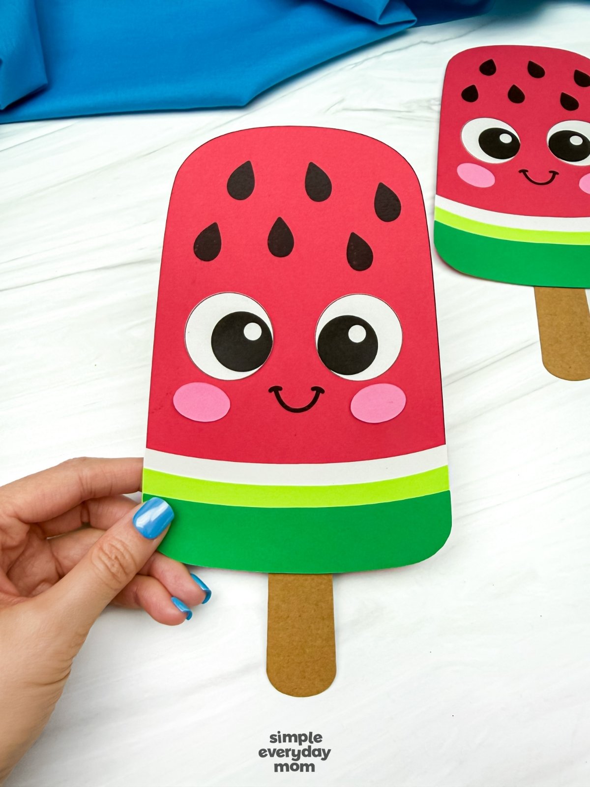 Hands holding a completed summer popsicle craft with a smiling watermelon face and popsicle stick handle on a white tabletop.