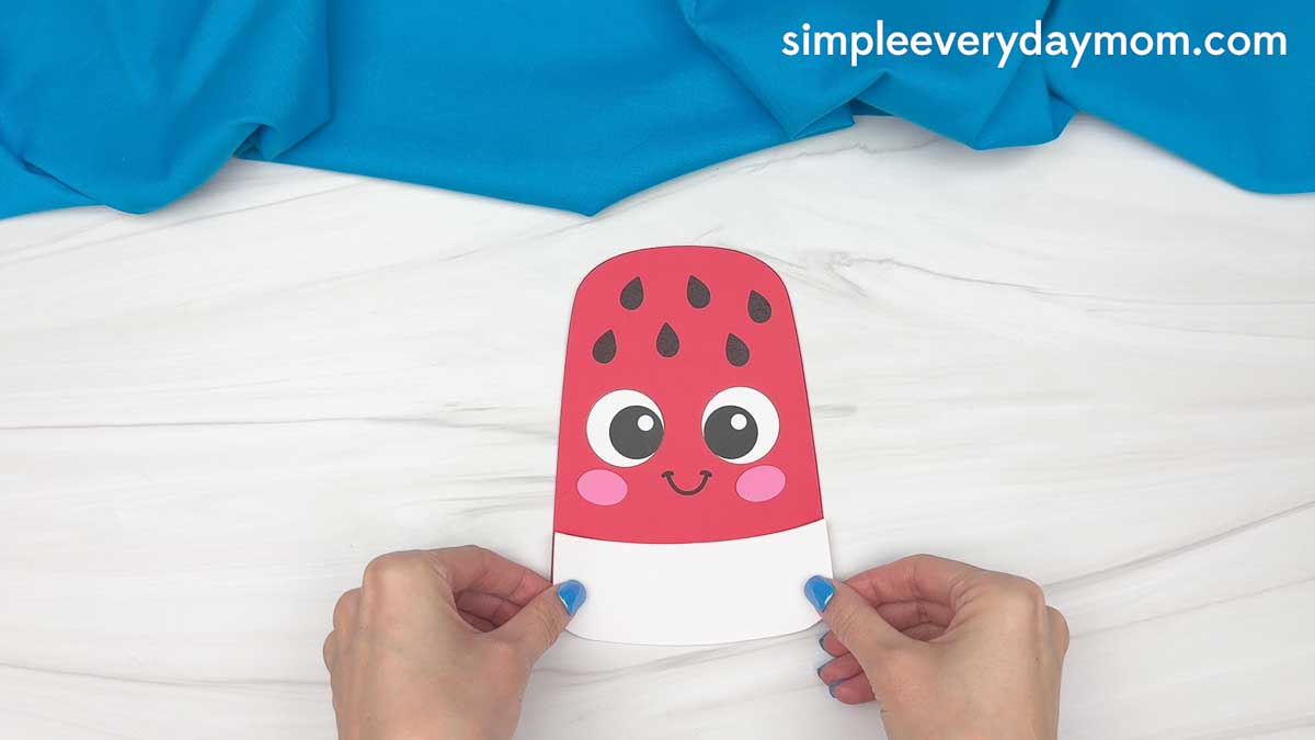 Hands holding a completed summer popsicle craft with a smiling watermelon face and white bottom section before adding the final stripe.