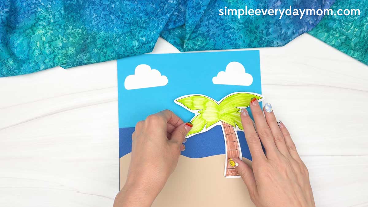 Hands attaching a colored palm tree cutout to a beach background for a reindeer summer craft.