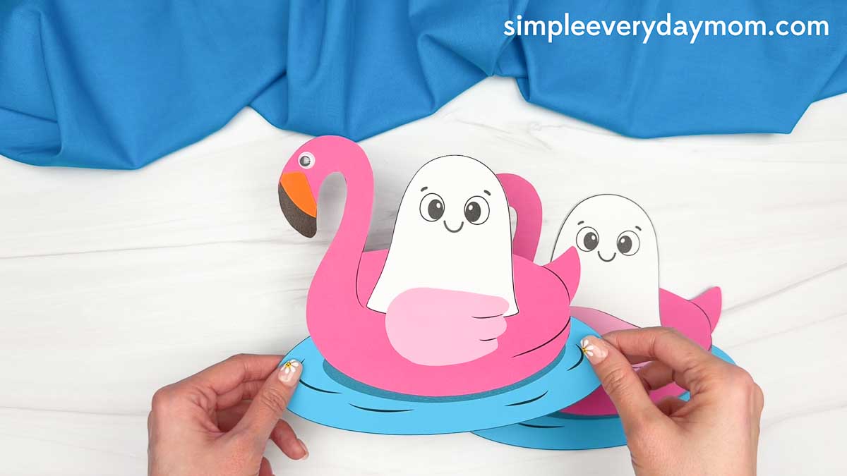 Hands holding a completed Summerween ghost craft with paper ghosts floating in pink flamingo pool floats.