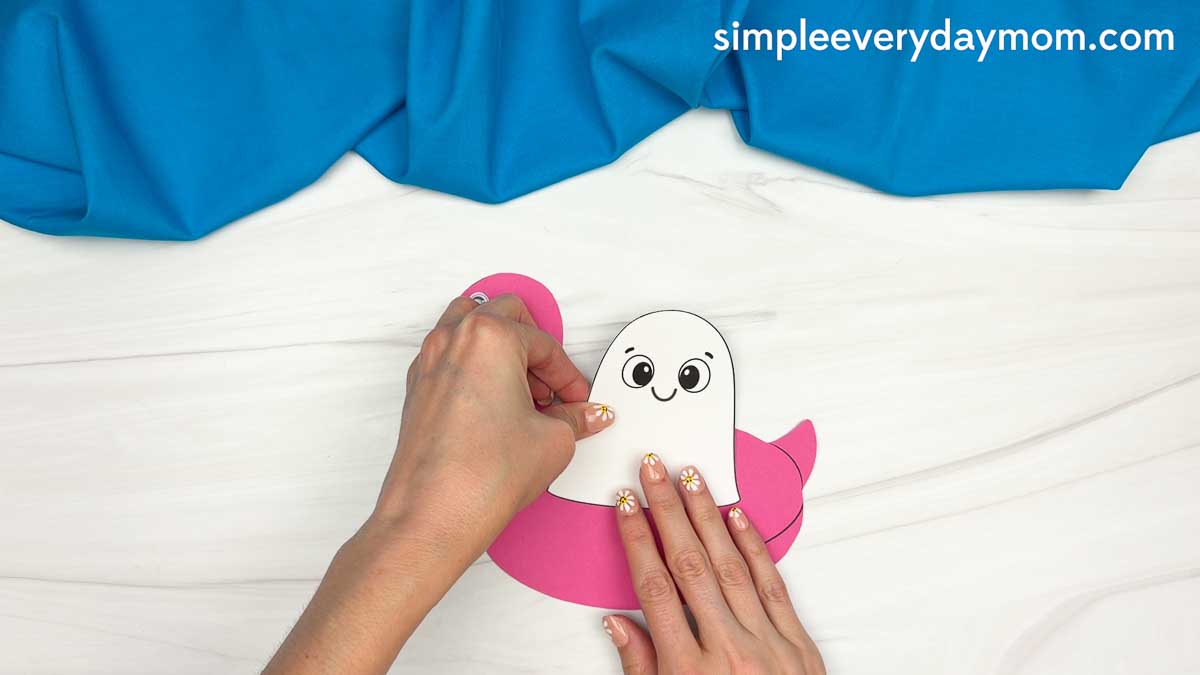 Hands attaching the ghost cutout to the pink flamingo pool float for the Summerween ghost craft.