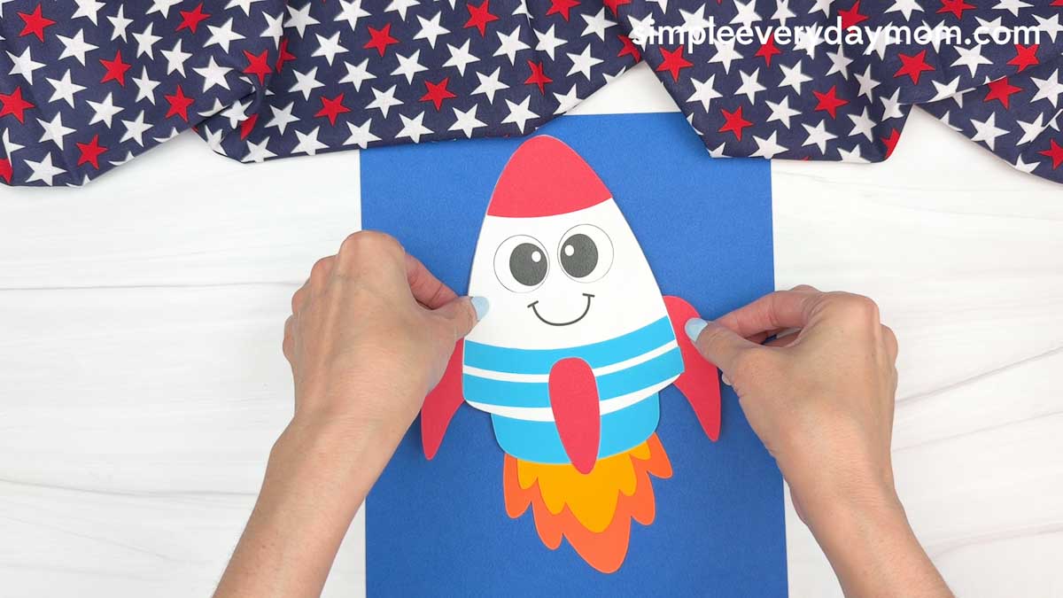 Hands placing a completed 4th of July rocket craft onto blue cardstock to create a patriotic paper craft scene.
