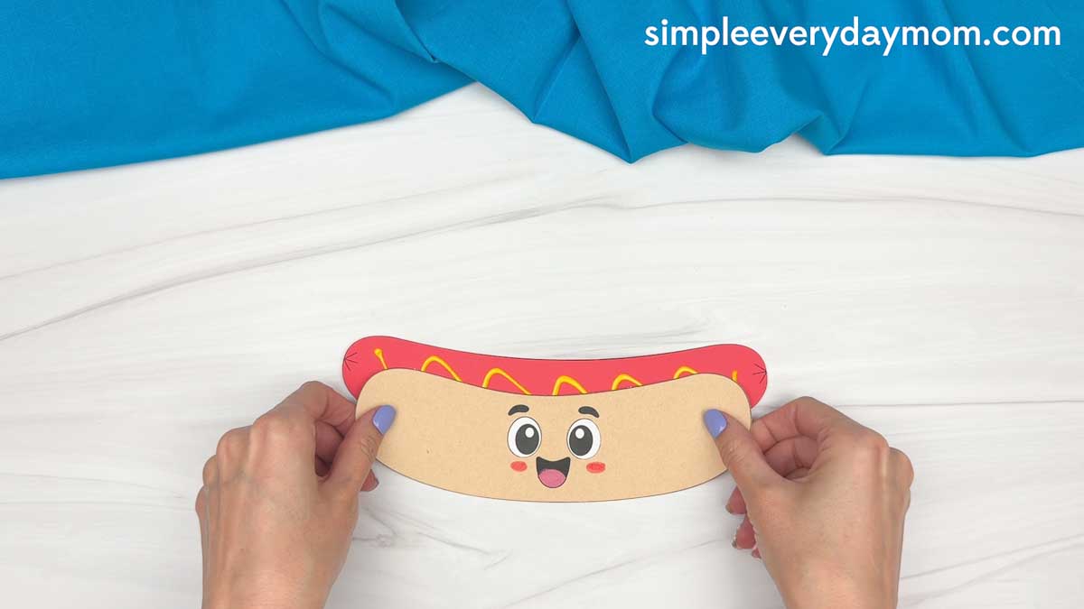 Hands gluing the smiling paper bun underneath the hot dog cutout to assemble the hot dog craft.