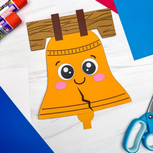 A completed Craft featuring a smiling Liberty Bell paper craft surrounded by colorful construction paper, glue sticks, and scissors.