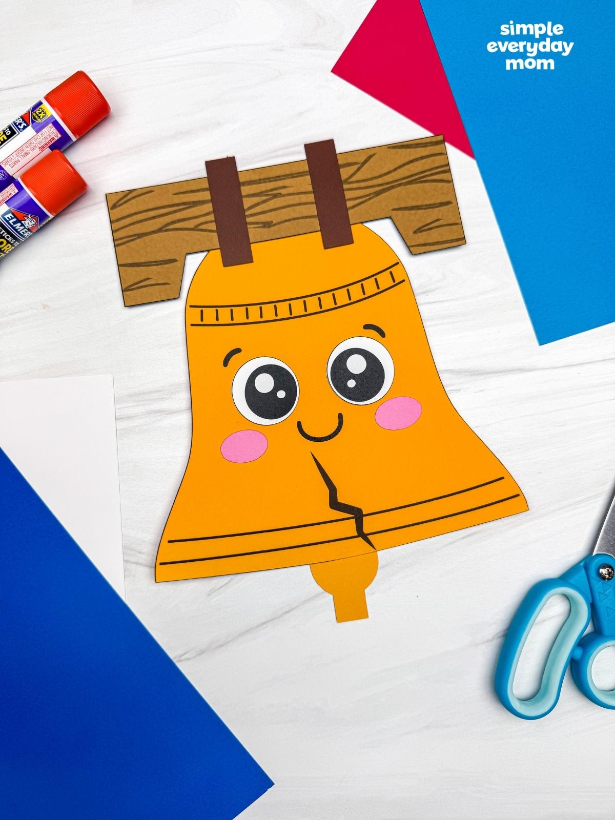A completed Craft featuring a smiling Liberty Bell paper craft surrounded by colorful construction paper, glue sticks, and scissors.