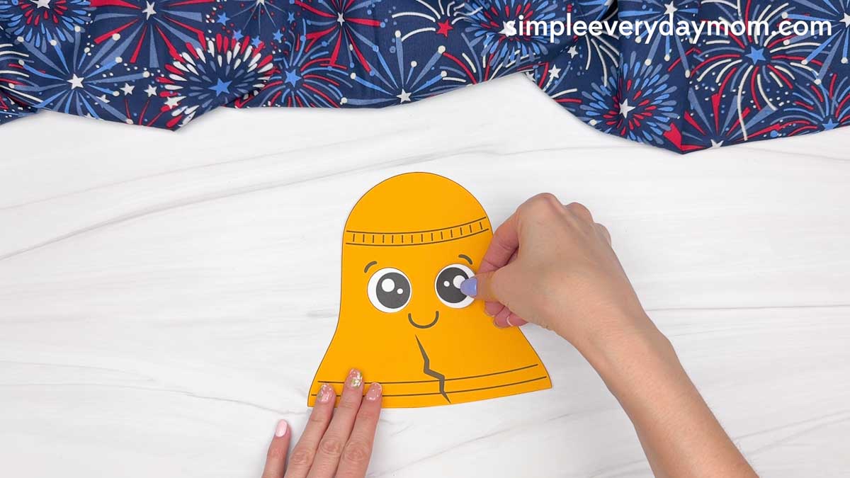 Hands placing an eye piece onto a cut out Liberty Bell paper craft during assembly.