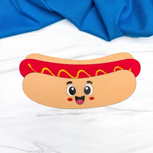 A completed Craft showing a smiling paper hot dog craft with mustard topping on a white tabletop.