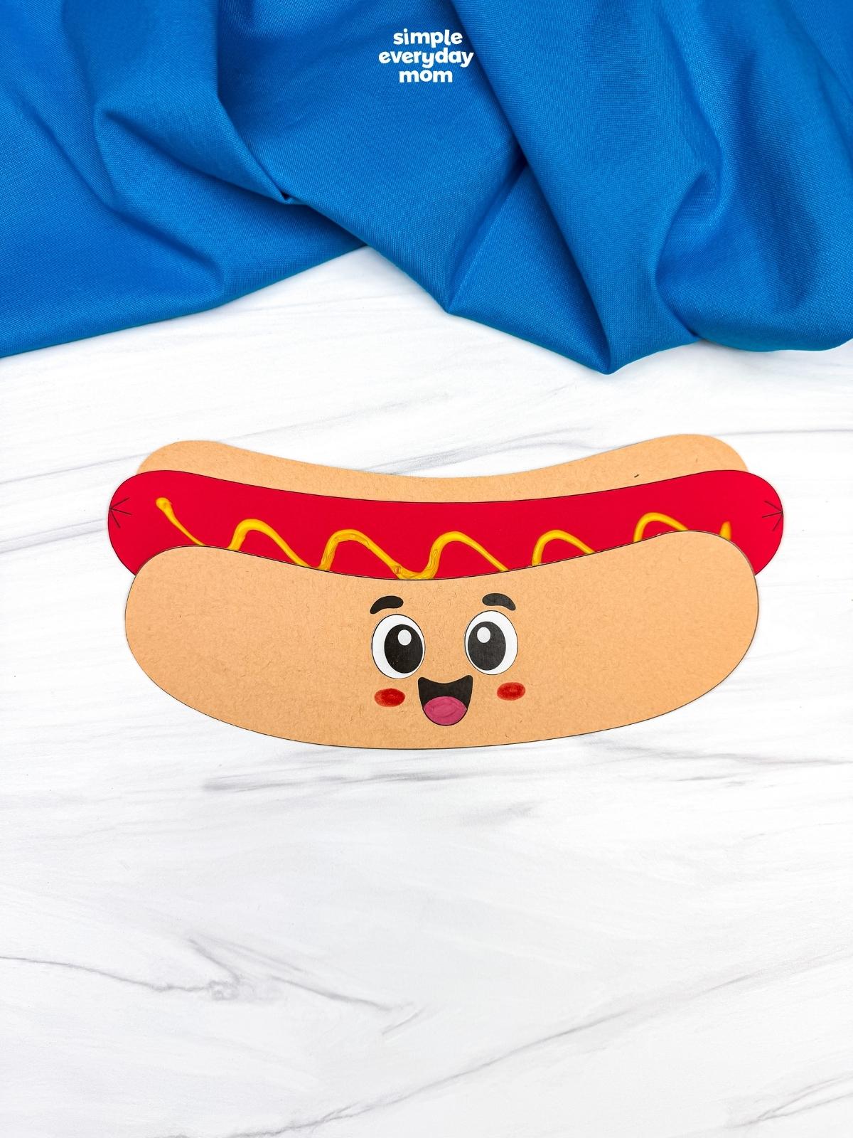 A completed Craft showing a smiling paper hot dog craft with mustard topping on a white tabletop.