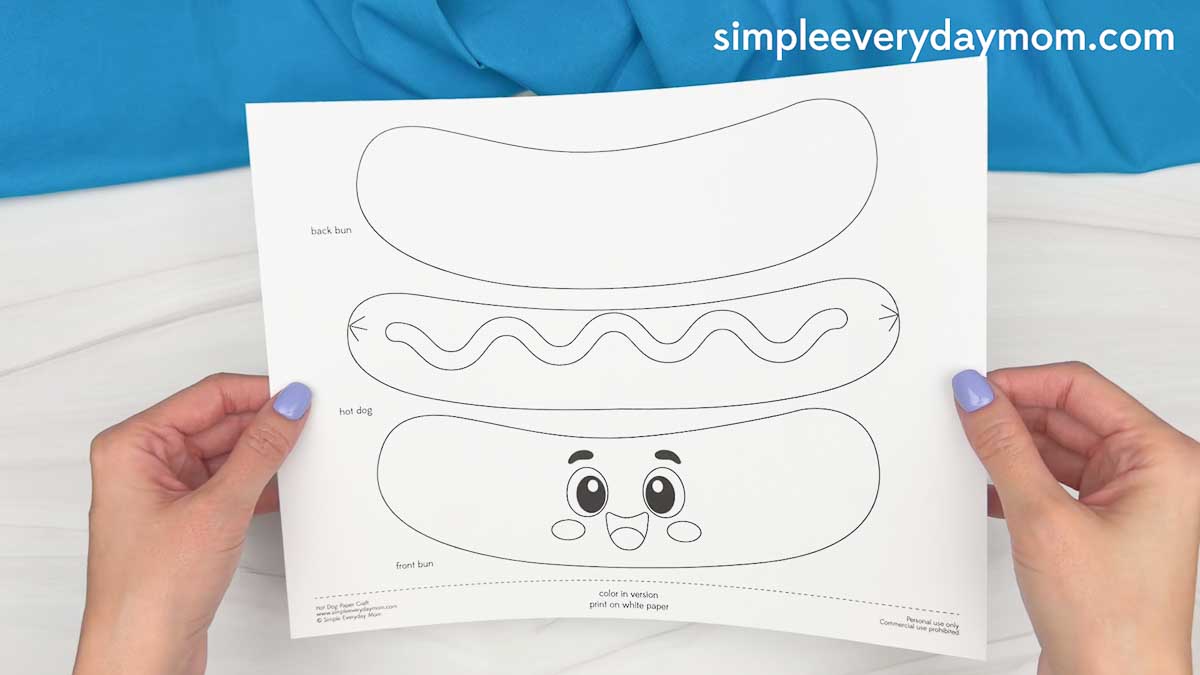 Hands holding a printed hot dog craft template with bun and hot dog cutout pieces on a sheet of paper.