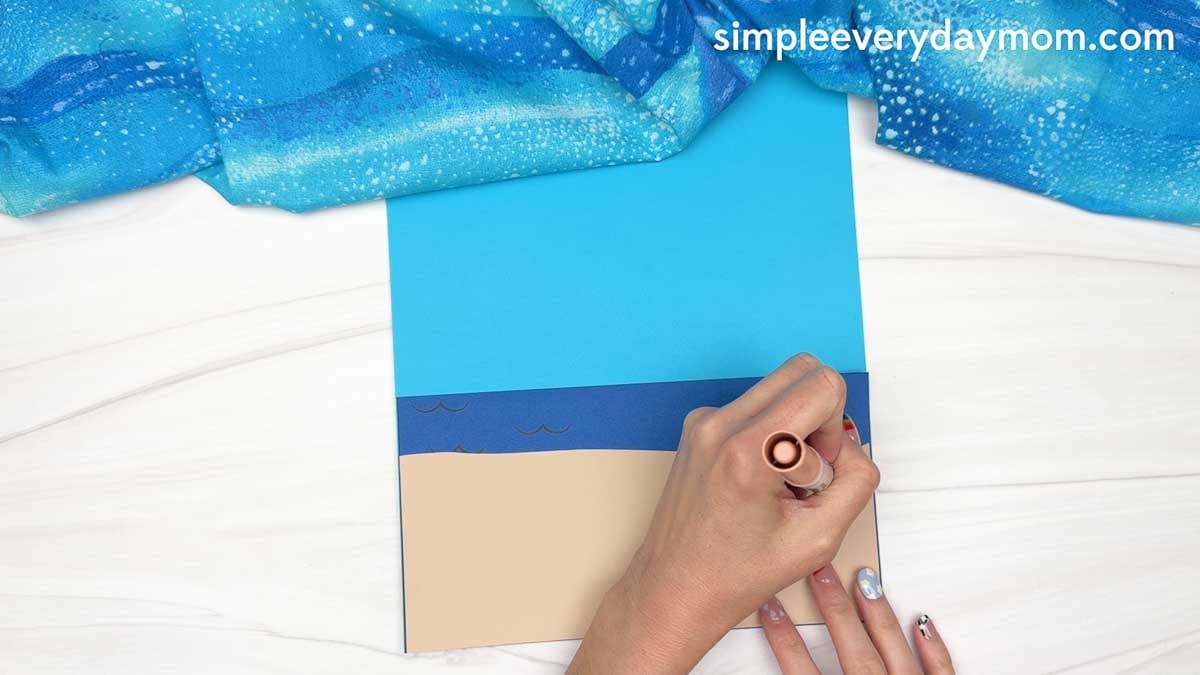 Hands draw details on a beach scene background made with blue ocean, sky, and sandy shore cardstock for a Christmas in July gingerbread man craft.