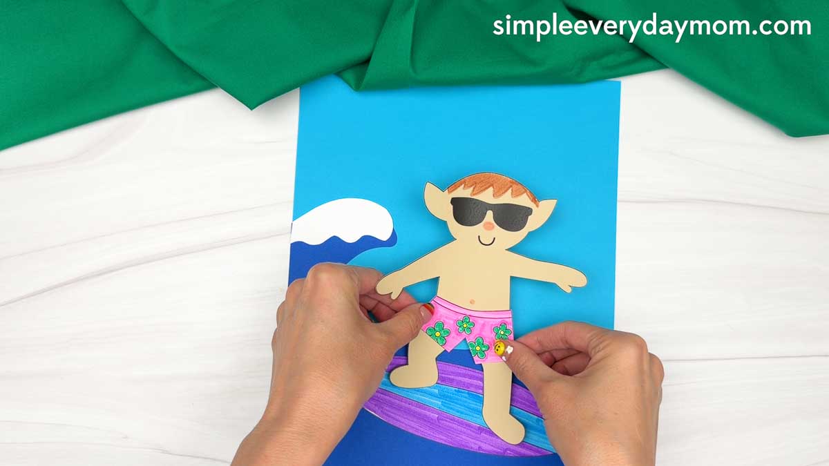 Hands placing a completed elf cutout onto a colorful surfboard against an ocean wave background for an elf surfing craft.
